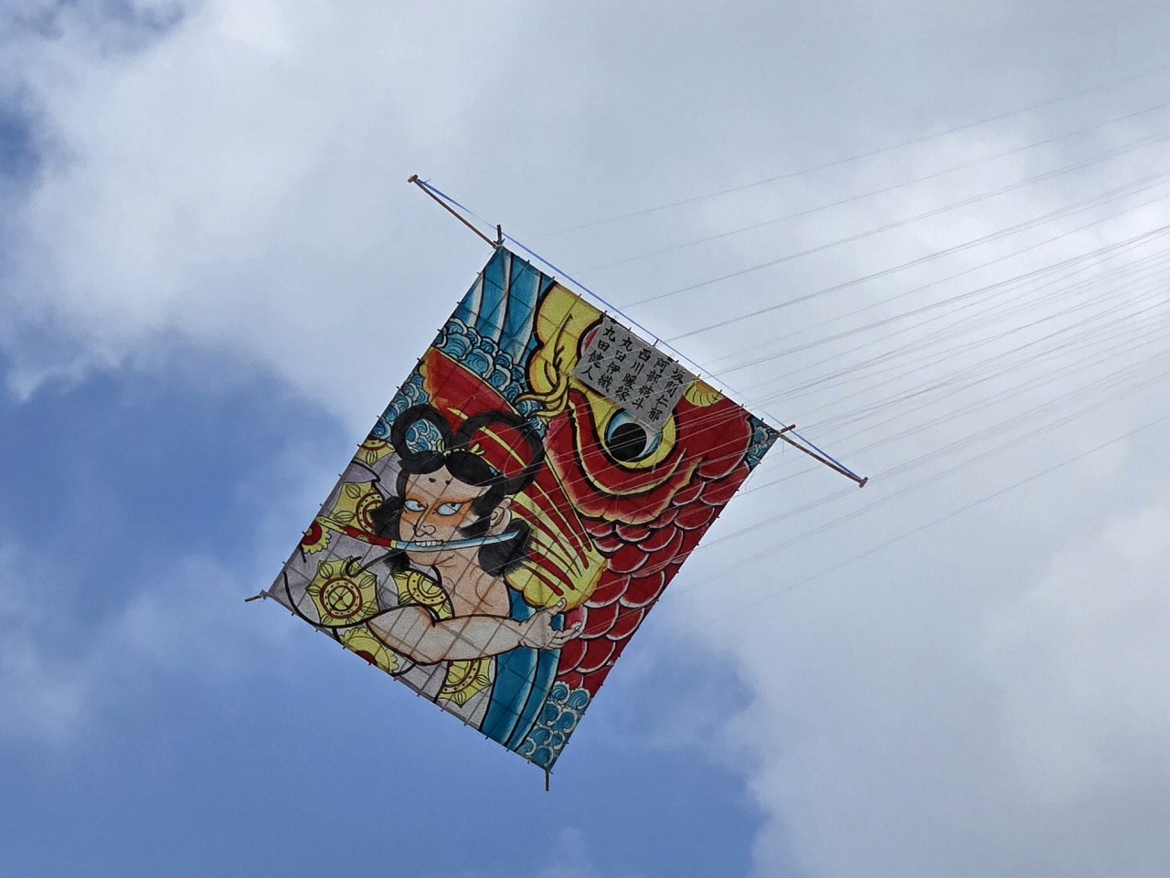 日本の和柄のイラストが描かれた、赤い鯉と刀をくわえた人物の模様の大きな凧が、複数の凧糸で空に高く揚がっている写真