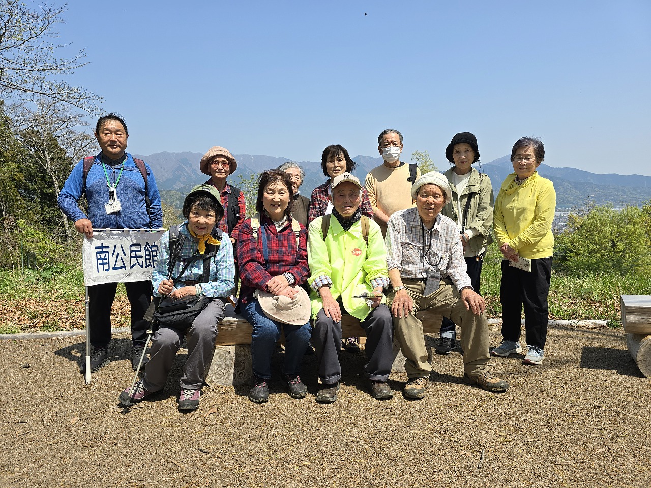 快晴の空の下、山々の景色を背景に、ハイキングを楽しむ複数の参加者が「南公民館」と書かれた旗を持ち、ベンチに座ったり立ったりしながら、集合写真を撮影している様子の写真