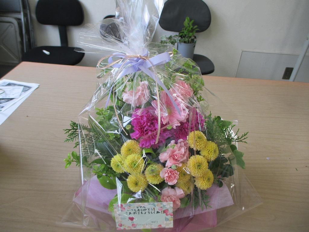 透明なフィルムでラッピングされ、リボンが結ばれた花束がテーブルに置かれており、「ママいつもありがとうこれからもよろしく」と書かれたメッセージカードが添えられている写真
