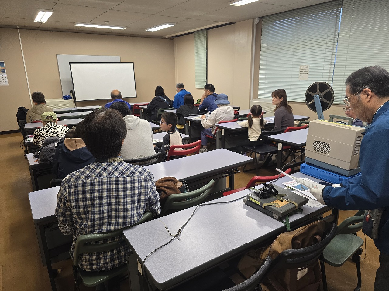 複数の参加者が机と椅子に座って前方にあるスクリーンの方を向き、右手前に機械を操作する男性の写真