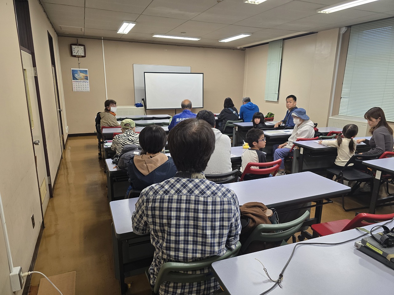 複数の人々が教室で机と椅子に座り、前方にあるスクリーンに向かって座っている様子を、後方から捉えた写真