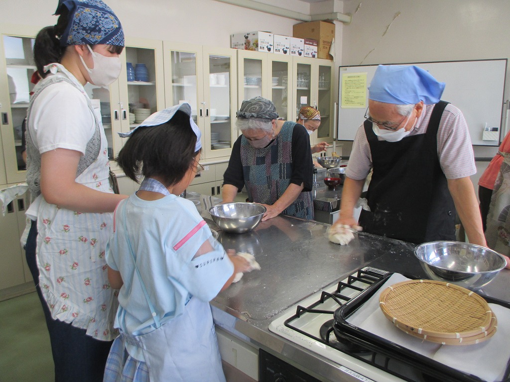 参加者がエプロンや三角巾、マスクを着けて厨房の調理台を囲んで、それぞれ手作業でこねたりしている様子の写真