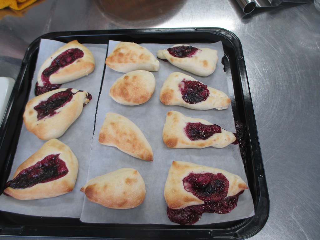 天板に敷かれたクッキングシートの上に、紫色のベリーソースがはみ出したパンが複数並べられており、焼き立ての熱気が伝わってくるような出来栄えの写真