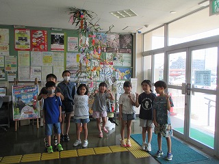 たくさんの笹飾りが天井から吊るされた室内で、数人の子供たちがカメラに向かってポーズを取り、楽しそうな笑顔を見せている様子の写真