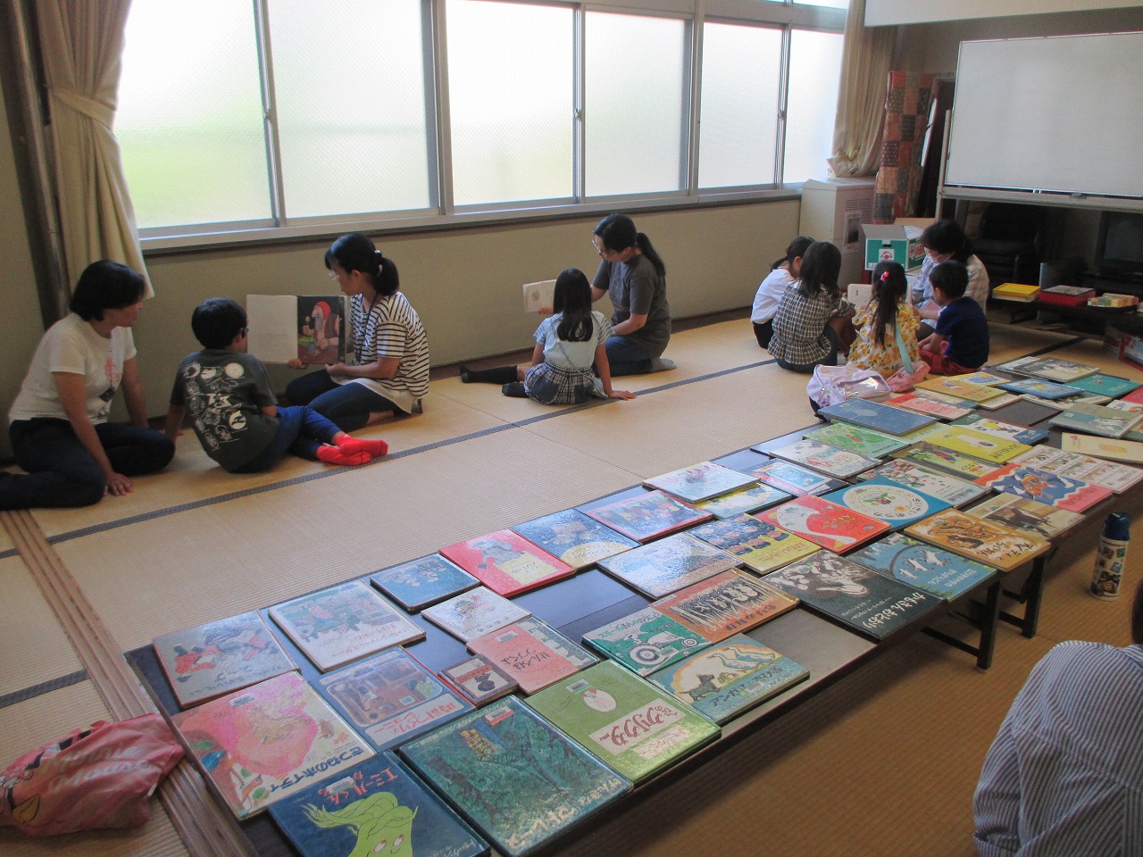大きな和室で、複数の親子が畳の上に座り、床に広げられた多数の絵本の中から、それぞれ好きな本を手に取って読んでいる様子の写真