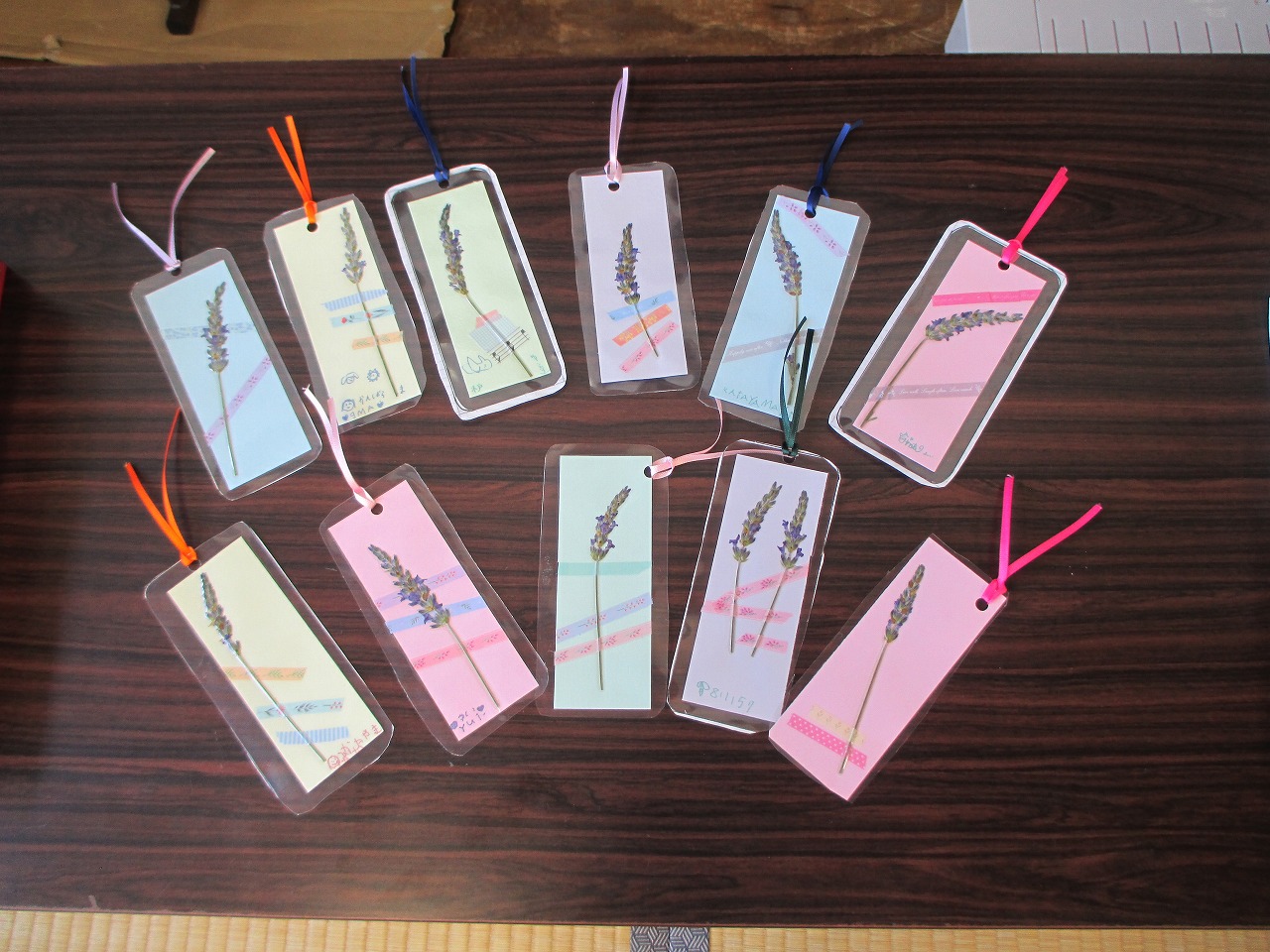 様々な色の紙にドライフラワーやマスキングテープを使い、ラミネート加工された複数のしおりが、それぞれ異なる色のひもを付けてテーブルの上に並べられている写真