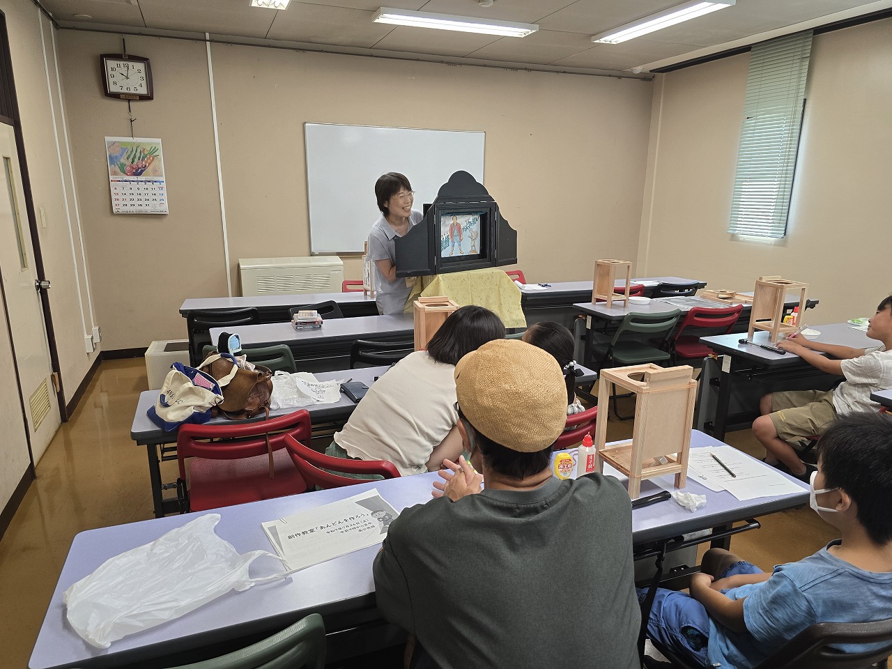 教室内で机に座った子どもたちと保護者が、前方で紙芝居を演じる女性の話に集中して耳を傾け、各机には木製の工作キットが置かれている写真