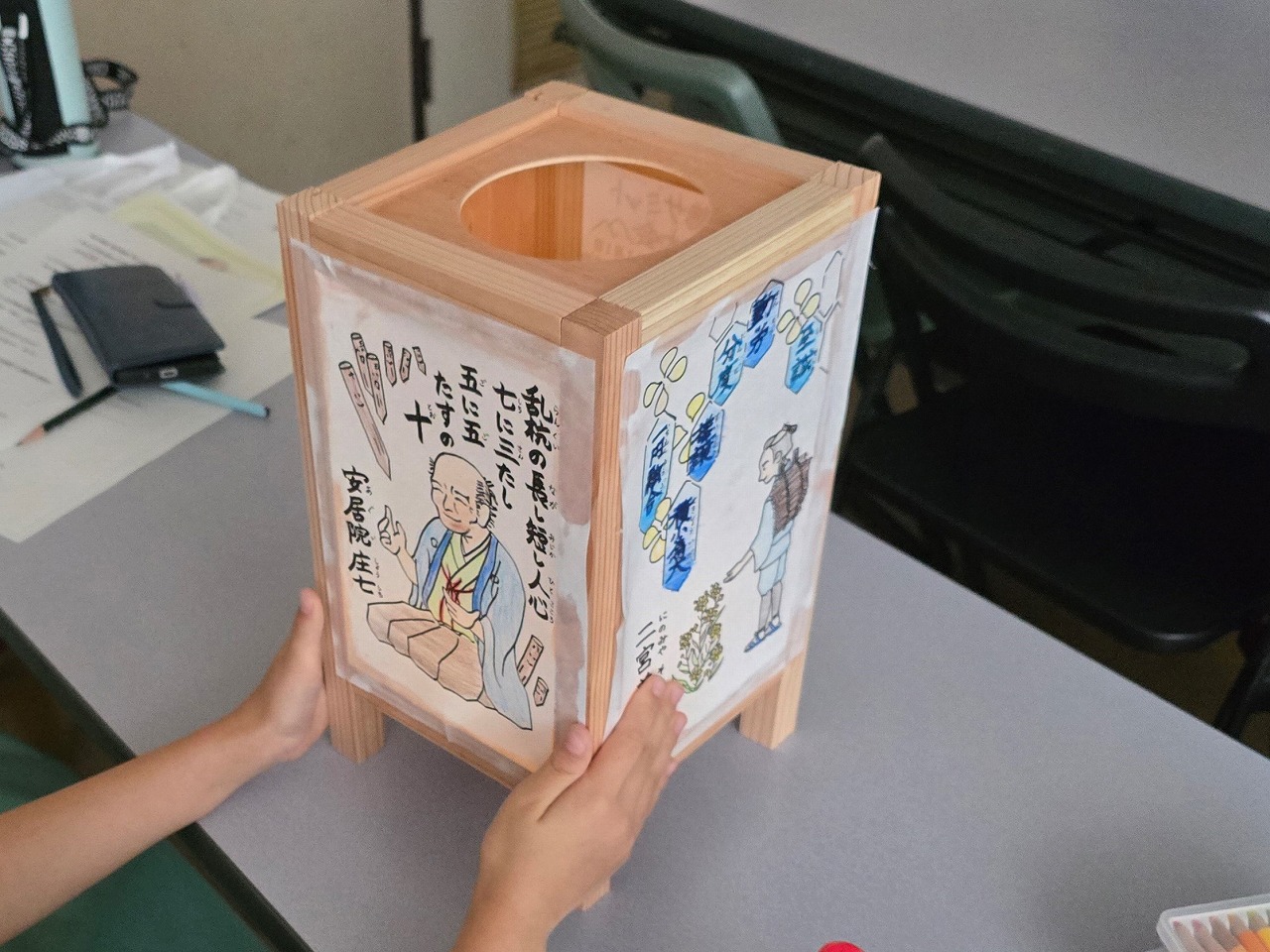 机の上で両手で抱えられている、木枠に手描きでイラストや文字が描かれた、和風デザインの四角い灯籠の写真