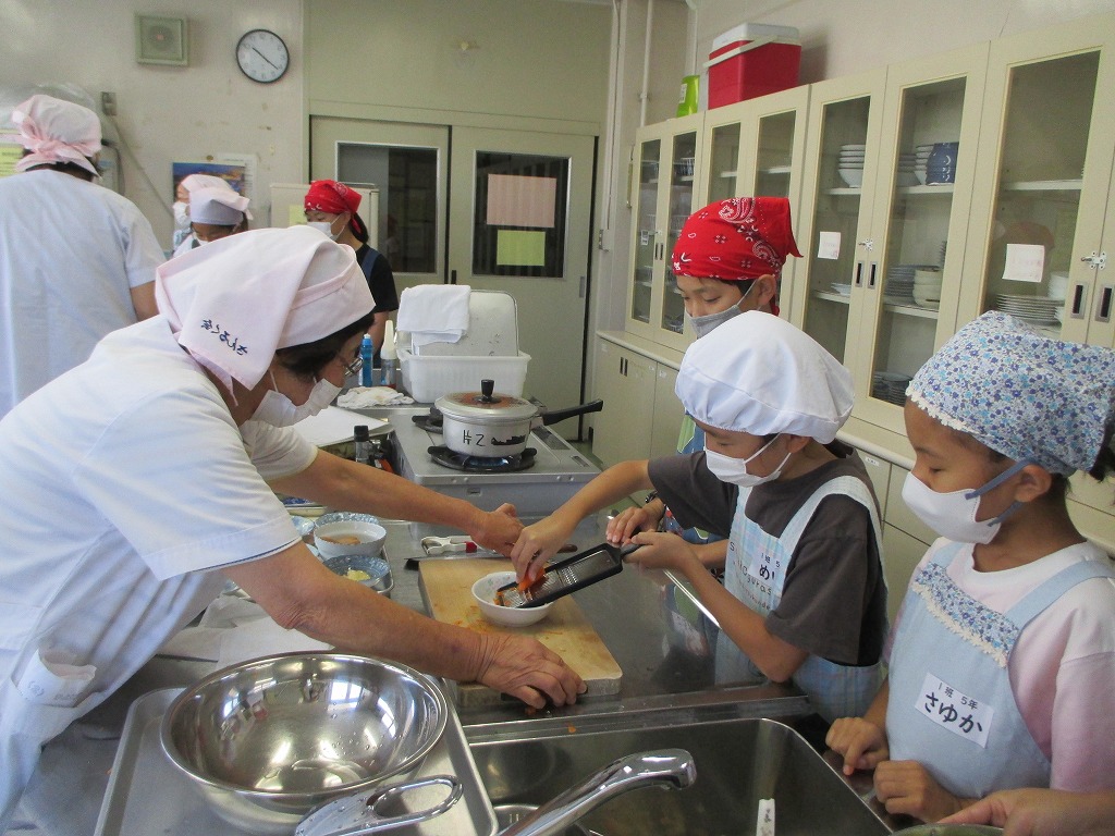 子どもたちがエプロンと三角巾、マスクを着用し、厨房で大人の指導を受けながら、野菜をすりおろすなどの調理作業に真剣に取り組んでいる様子の写真