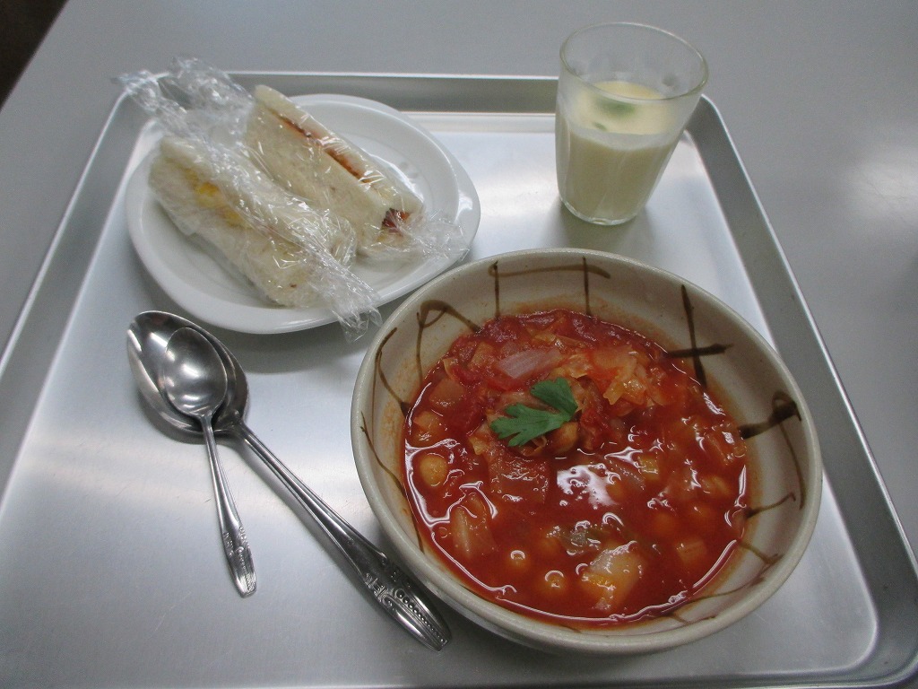 銀色のトレーの上に置かれた、ラップに包まれたサンドイッチ、大きなボウルに入ったひよこ豆とキャベツのトマトスープ、飲み物の写真