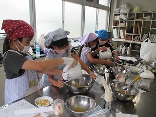 子供たちがマスクとエプロンを着用し、ボウルやハンドミキサーを使って熱心に菓子作りをしている様子の写真