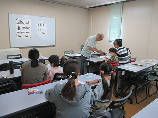 教室の机に座った子どもたちとその保護者が講師の説明を聞きながら、昆虫標本や模型を使った学習活動に取り組んでいる様子を後方から撮影した写真