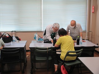 高齢の男性ボランティアが机に座る女性の壊れた品物を確認しながら修理を行っている様子を写した写真