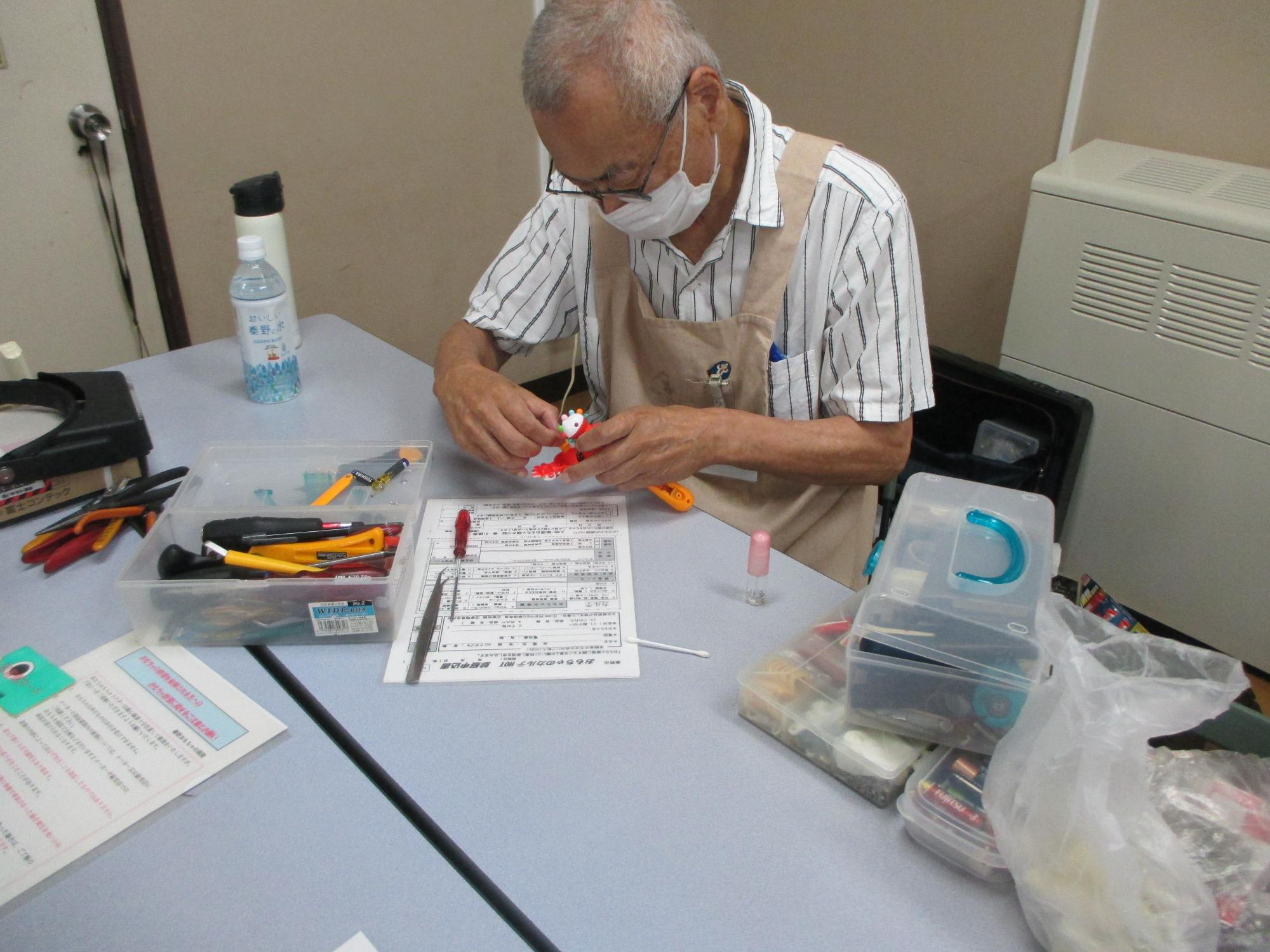 作業台に工具や修理用の道具が広げられた中で、男性ボランティアがおもちゃを丁寧に分解・修理している様子の写真