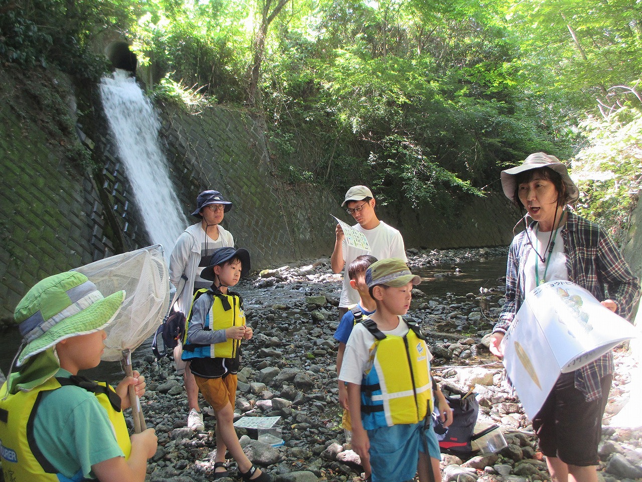 複数の子供と大人が川辺に集まり、帽子やライフジャケットを着用した子供たちが網を持ち、大人の指導者が何かを説明している様子の写真