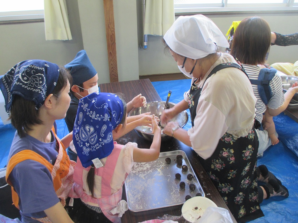 親子が三角巾とエプロンを着けてテーブルを囲み、講師と一緒に大きなボウルで生地をこねながら団子を作っている様子の写真
