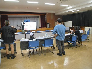 会議室のような広間で、若い講師の男性がプロジェクターの画面を指し示し、数名の聴講者に対してワークショップ形式の講座を行っている様子の写真