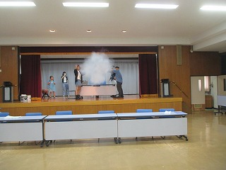 室内の舞台の上で男性が大量の白い煙を発生させ、数名の子どもや関係者が見守っている科学実験イベントの様子の写真