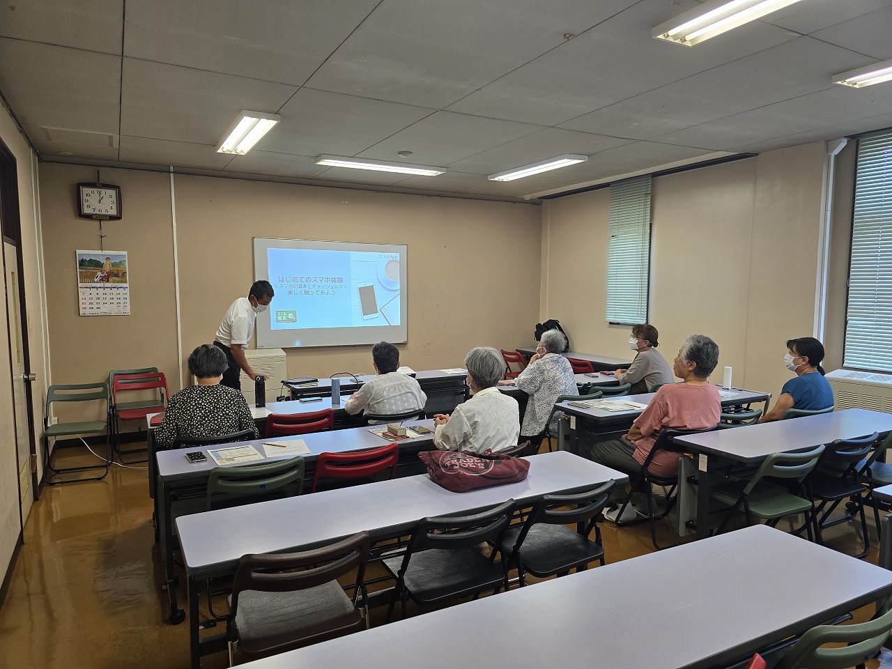 教室で、講師の男性がプロジェクターに投影された「はじめてのスマホ体験講座」の資料を指し示し、数名の高齢者が机に向かい熱心に受講している様子の写真