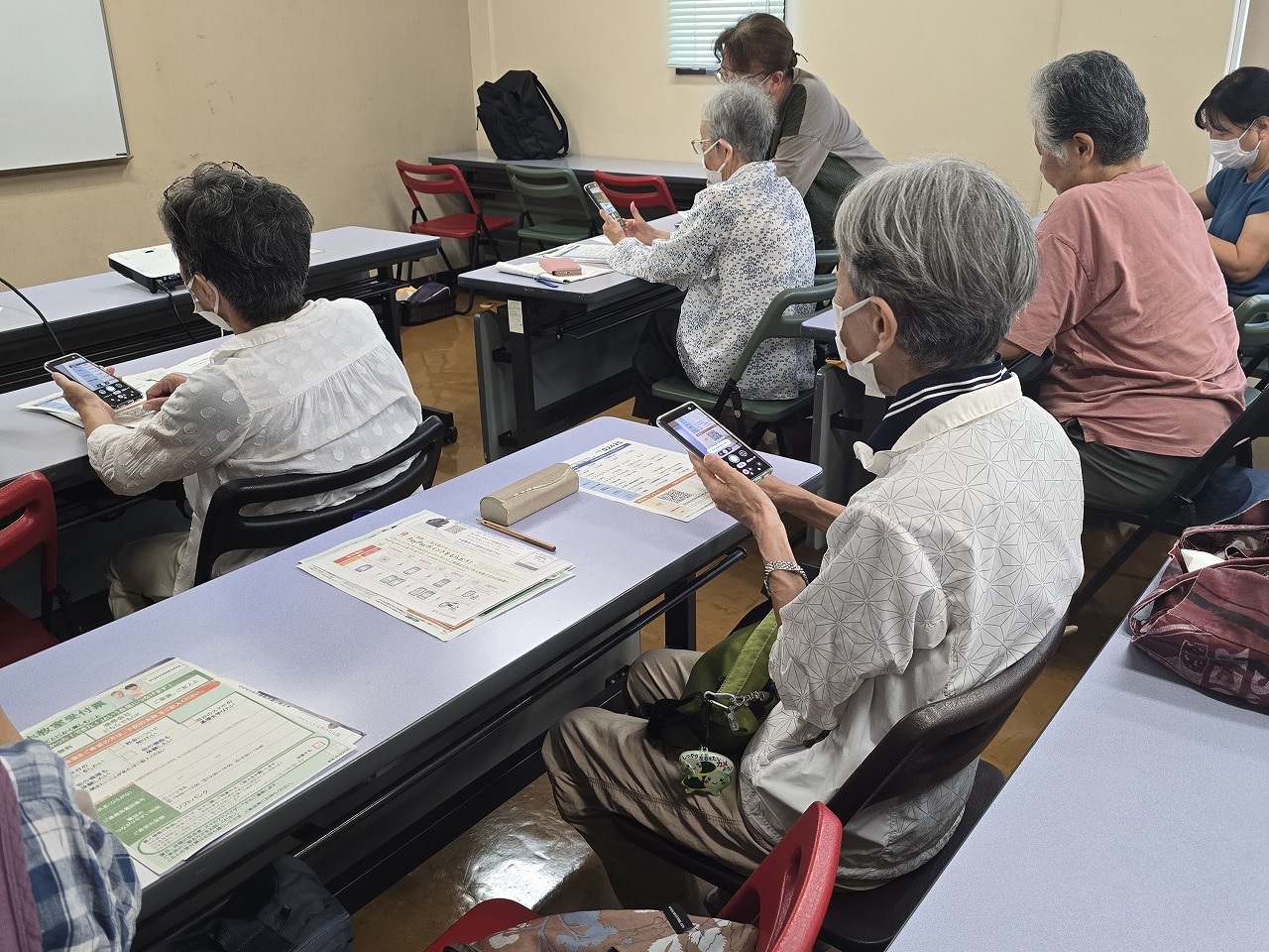 教室内で、マスクを着用した数名の高齢者の女性が、机の上に置かれた資料を見ながら、スマートフォンを操作して実習に取り組んでいる様子の写真