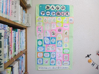 木製の本棚が並ぶ図書館の壁に、淡い緑色の台紙に「みんなのアートぬりえ」と書かれたタイトルと、複数の小さな塗り絵の作品が展示されている写真
