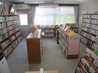 窓からの光が白いカーテンを通して入る明るい室内で、天井まで届く木製の書棚と背の低い陳列棚が複数並び、たくさんの本が整理されて収納されている図書室の写真