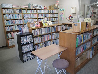 木製の高い本棚と低い陳列棚がコの字型に配置され、たくさんの本が並べられた図書館または図書室の室内で、手前には一人掛けの折りたたみ机と丸椅子が置かれている写真