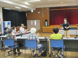 室内で、女性の講師が立ち、椅子に座った数名の高齢者が拍手をしている様子を捉えた写真