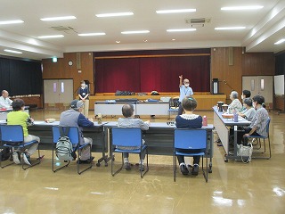 大会議室のような広間で、講師の男性が聴講している十数名の高齢者に対し、壇上で身振り手振りで何かを説明している様子の写真