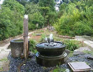 奥には草木が生い茂り、手前には玉砂利が敷き詰められた小さな石庭に設置された黒い鉢から水が噴き上がっている庭園の風景を写した写真