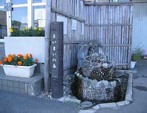 施設の入り口横に、「まいまいの泉」と縦書きされた木製の標柱が立ち、竹の塀を背景にして苔むした岩組の小さな井戸が設けられている写真