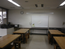 奥の壁際にホワイトボードが設置され、手前には木製の長机が左右に数台並んだ創作活動室の写真