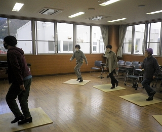 広い窓がある部屋で3人の参加者が、各自の白い板の上に立ち、講師の指示に合わせて手足を動かしている様子の写真