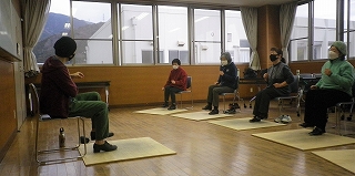 広い窓がある部屋で、参加者4名が椅子に座り、足元に敷かれた畳の上に足をのせ、講師の指示に合わせて胸の前で手を握り運動している写真
