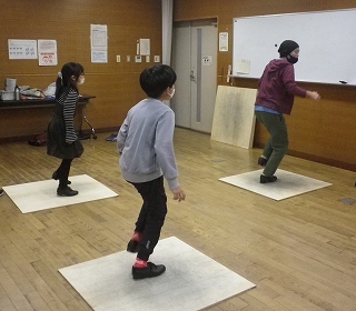 広い板張りの部屋で、大人一人と子供二人が、それぞれ足元に敷かれた白い板の上で、片足を上げてステップを踏んでいる写真