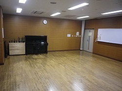 フローリング張りの室内左奥に黒いアップライトピアノが置かれ、入口横の壁にはホワイトボードが設置された音楽室の写真