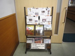 図書室の入り口付近に設置された木製のラックに、複数の新聞や雑誌が立てて展示されている写真