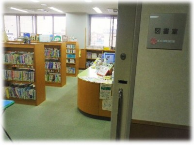 木製の書架に多くの本が並べられ、右側には角が丸い受付カウンターが配置されている、図書室の室内の様子を部屋の入り口から撮影した写真