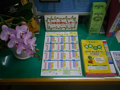 白い胡蝶蘭の鉢植えの横に、ゴミの分別収集カレンダーと「資源とごみ 分け方・出し方ガイド」の冊子が机の上に並べられている写真