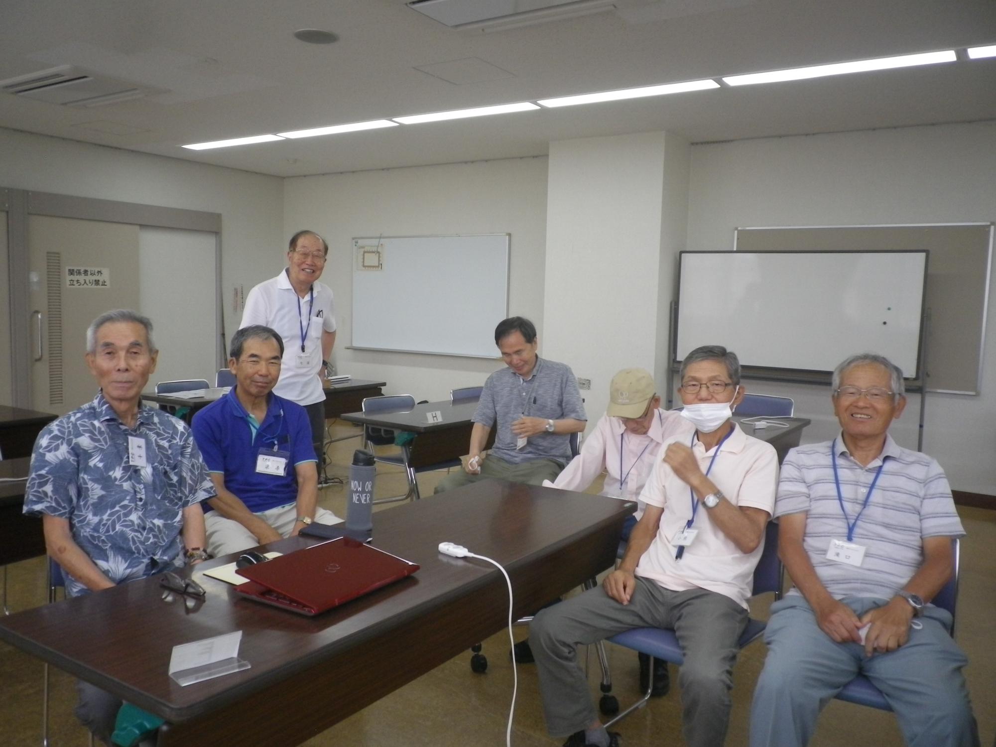 明るい室内で、複数の男性たちが長机を囲んで座り、笑顔で記念撮影している写真