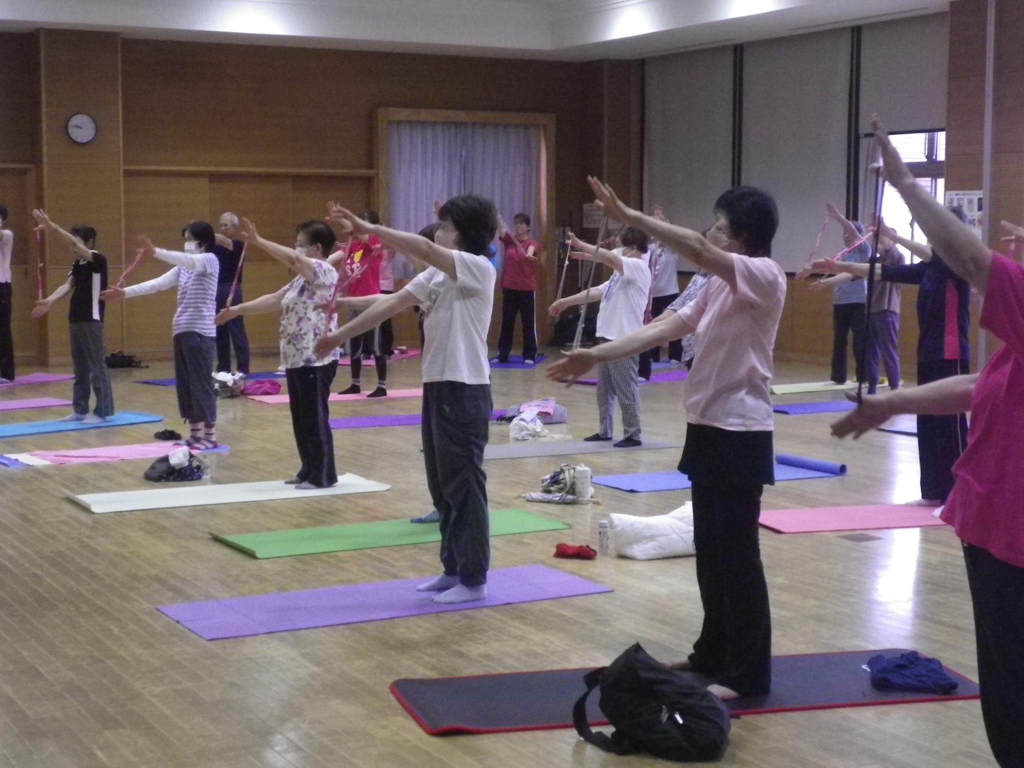 木製の床の広間で、大勢の人々がヨガマットを敷き、両腕を前または上に伸ばして体を動かしている様子の写真