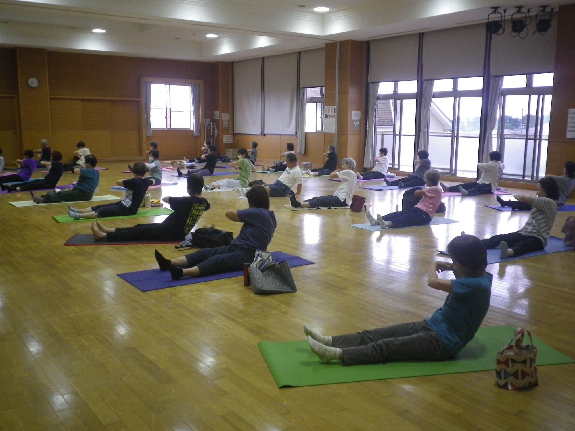 木目調の床の広間で、多くの参加者がヨガマットを敷いて床に座り、両足を前に伸ばし上半身を後方に傾ける動作をしている写真