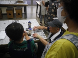 子供と高橋さんが、机に広げた新聞紙の上で、二宮金次郎像に熱心に着色しており、その後ろで女性が見守っている作業風景の写真