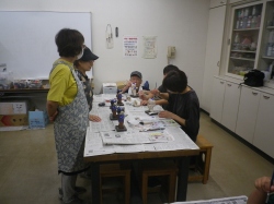 室内で複数の子供と大人が机を囲み、新聞紙を広げて二宮金次郎像に色を塗っており、後ろで別の大人が見守っている写真