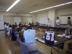 壇上で大人が何かを実演しながら説明しており、子供たちが机に座り真剣に聞いている様子の写真