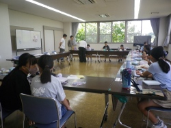 複数のテーブルをロの字型に配置した部屋で、子供たちが座って作業をしており、大人が指導しながら進行している様子の写真