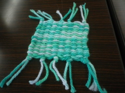 木目調のテーブルの上に置かれた、水色と白色の毛糸で手織りされた四角いコースターの写真