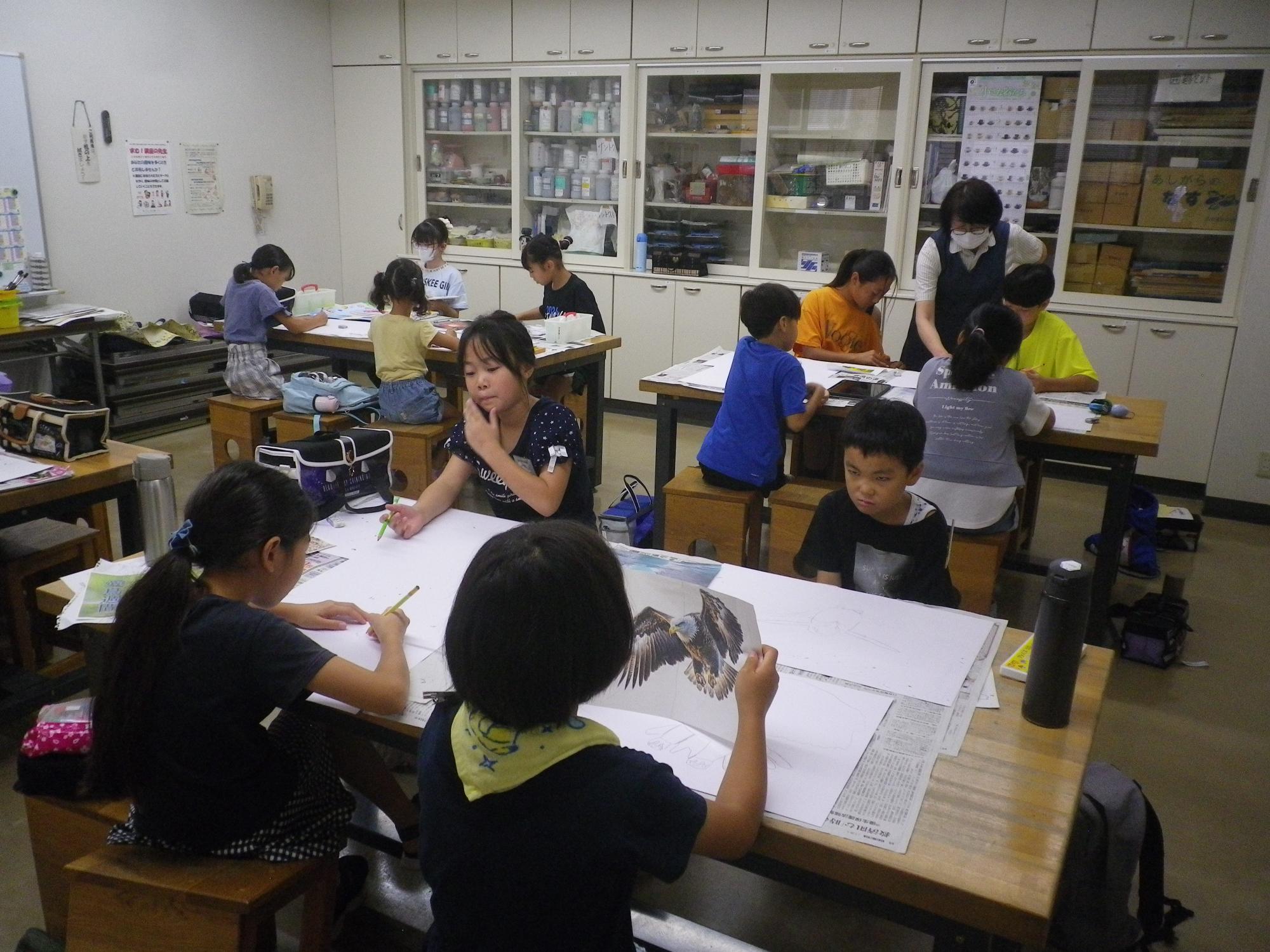 広い作業部屋の中で、子供たちが机に向かって絵を描いたり、紙を広げて作業をしたりしている写真