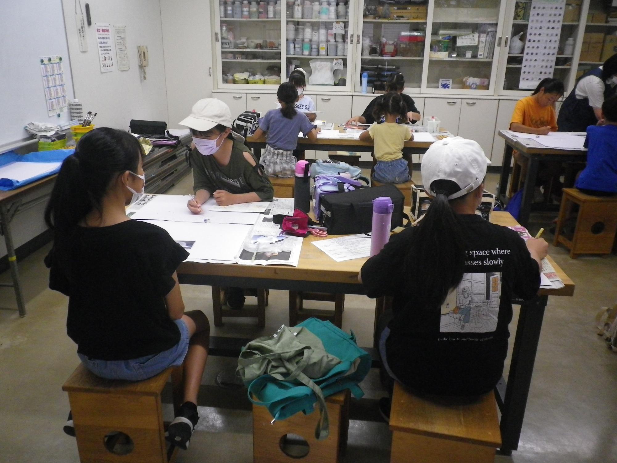 マスクをつけた子供たちが木製の椅子に座り、机に向かって絵を描いたり、紙を広げたりしている写真