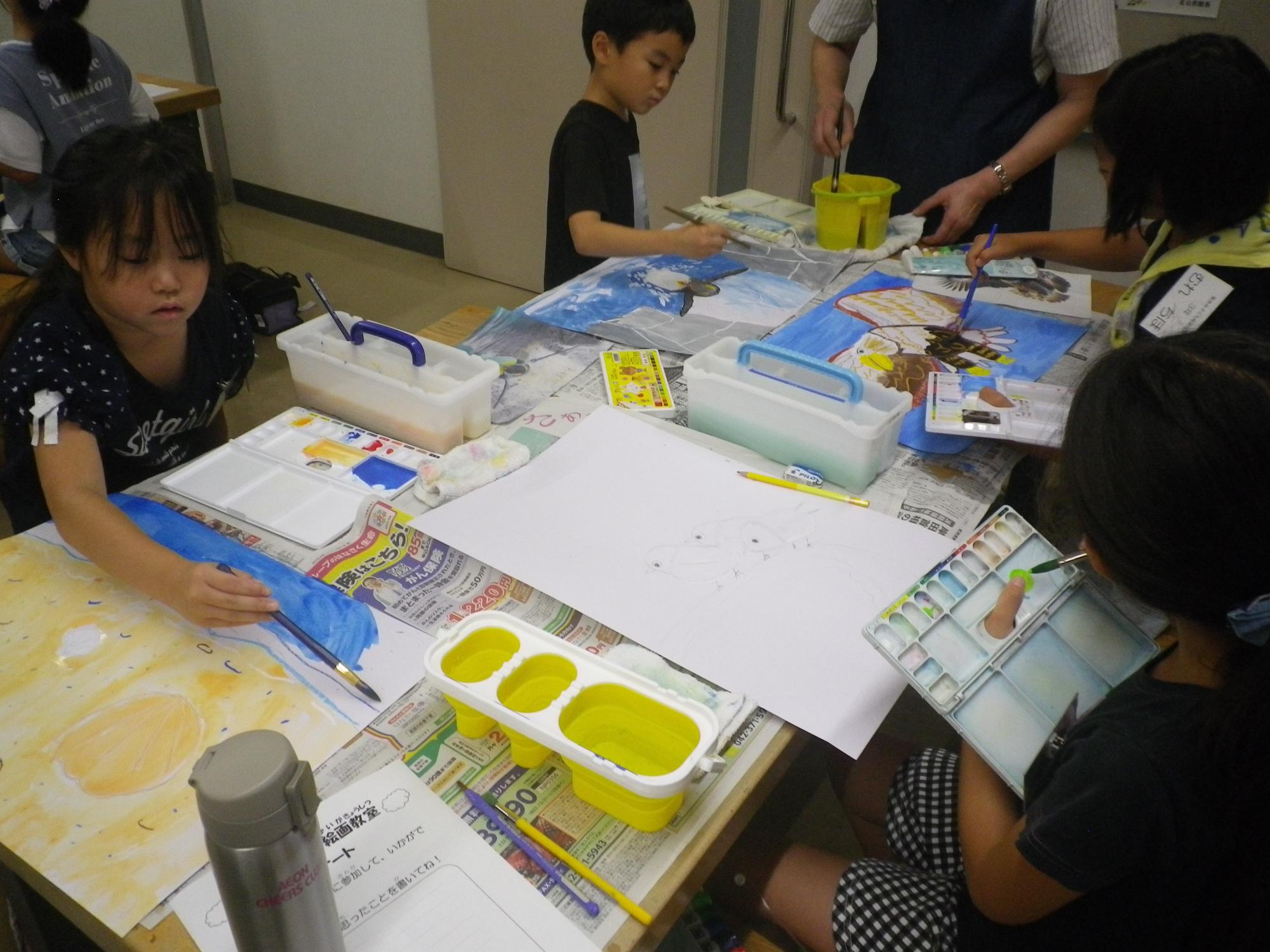 新聞紙を広げた机の上に水彩絵の具や筆が置かれ、子供たちが大きな紙に熱心に絵を描いている写真